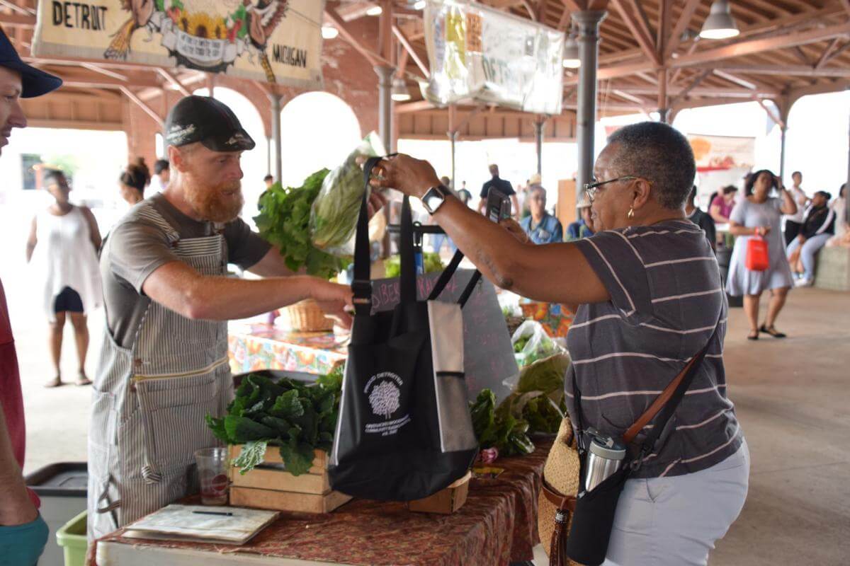 A man holding a bag up for a woman who is adding vegetables to her bag. | Long-Term Care Ombudsman Program