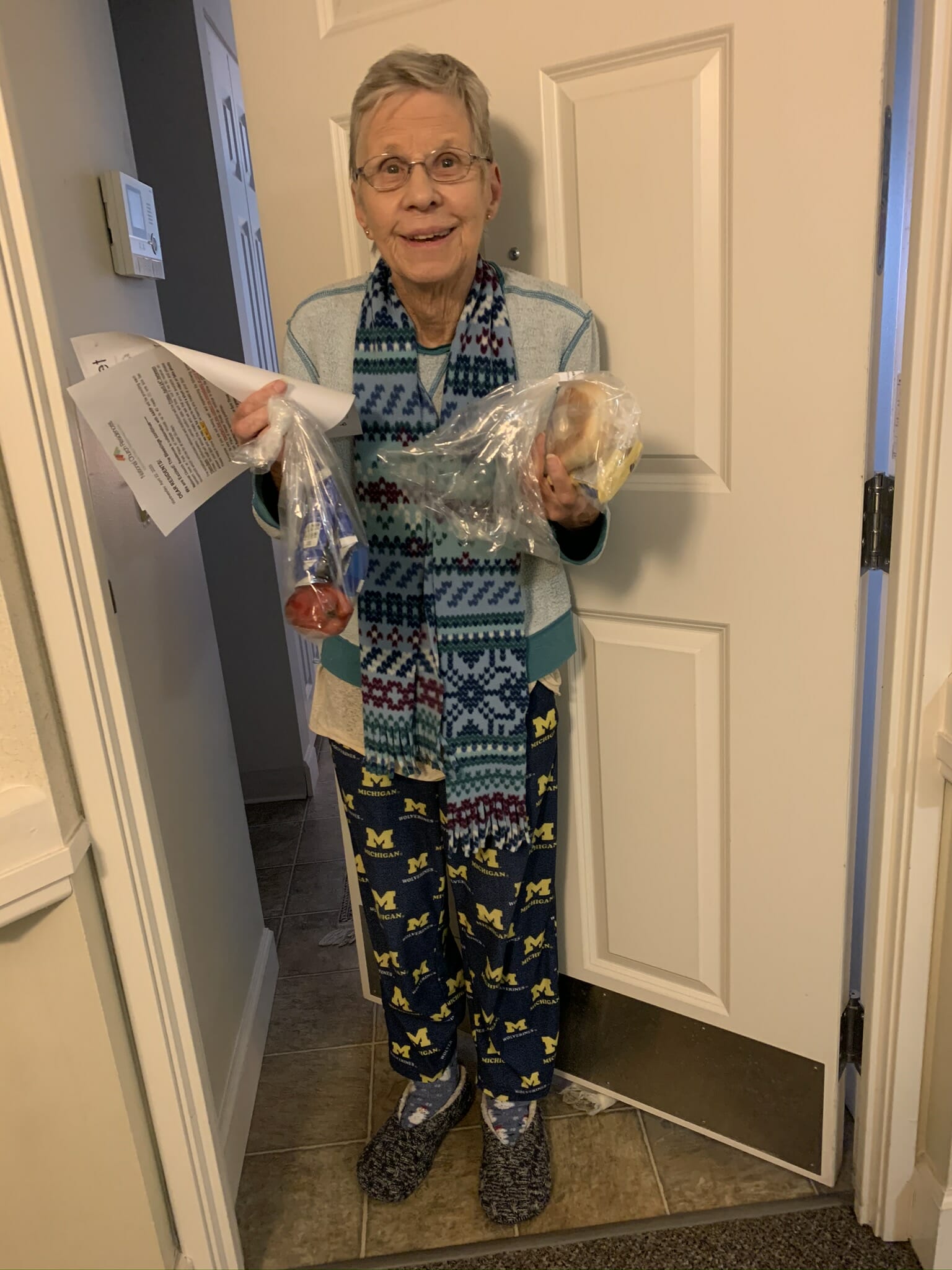 An older woman standing in front of her door holding papers and a bag with food